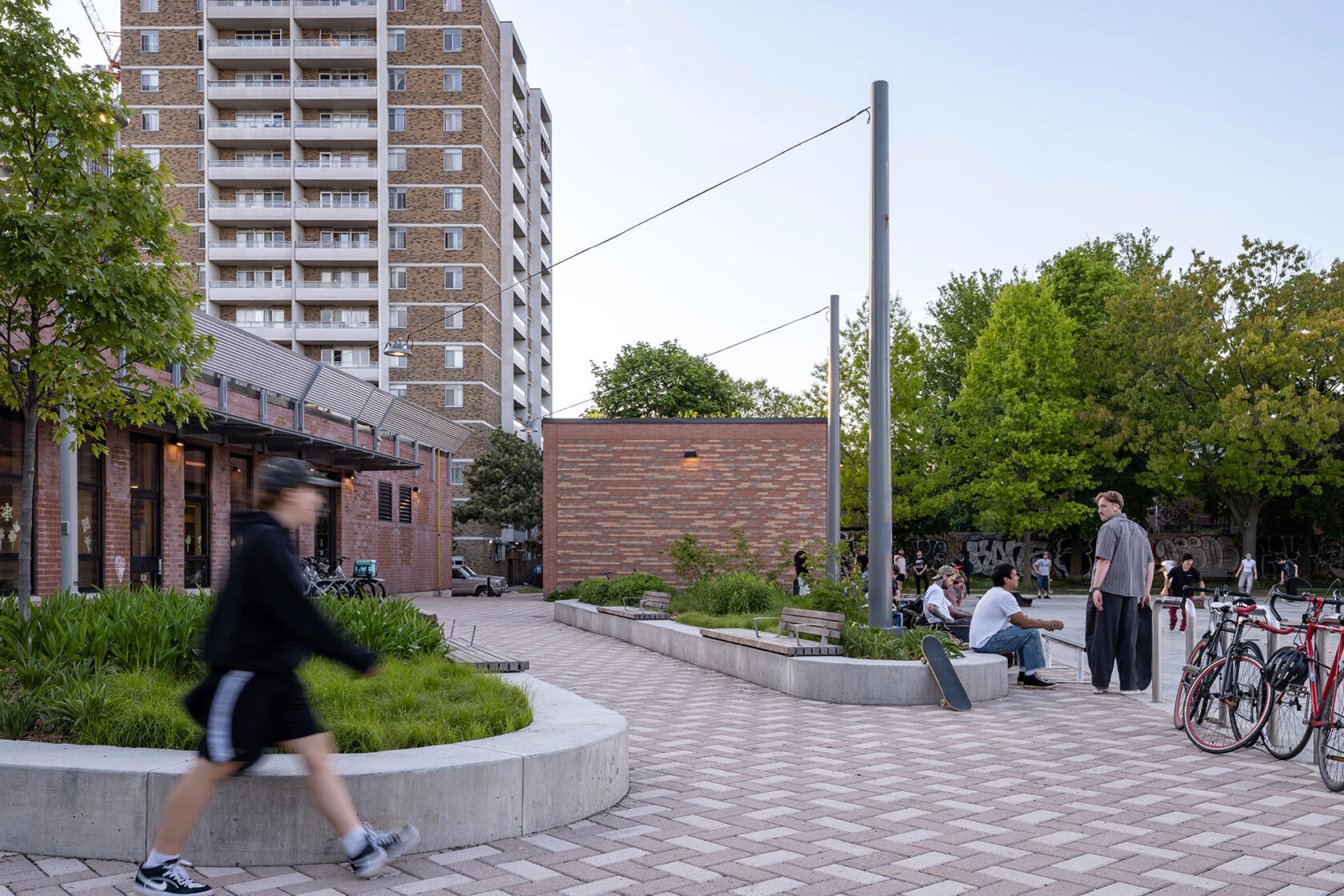 Low rise brick building with large windows and a large plaza with herring-bone paving pattern and planting lined with low seating walls. Next to the plaza is a skating rink. In the distance is a large apartment building