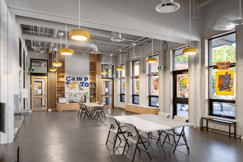 Interior of Dufferin Grove Clubhouse. Yellow pendant lights hang from a ceiling with exposed ductwork. Built in storage shelving and cupboards line the walls and the long rectangular windows look out on to the lively pedestrian plaza
