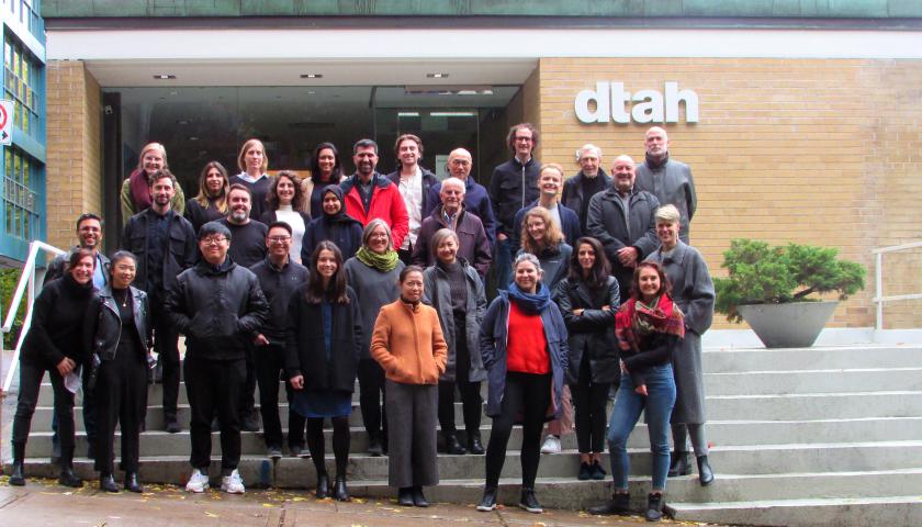 Group photo of people standing on steps in front of low rise brick building with DTAH sign beside glass front door.